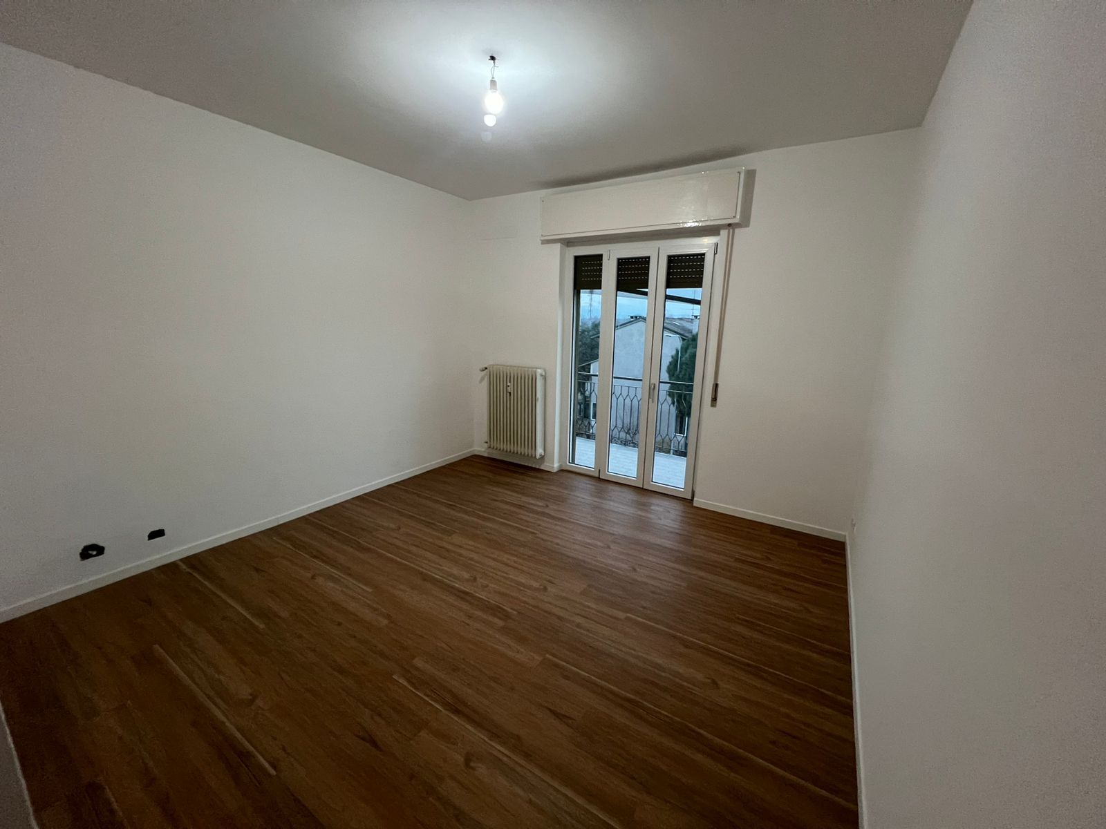 Stanza vuota con pavimento in legno, pareti bianche, lampada a soffitto, termosifone e porte finestre che danno sul balcone.