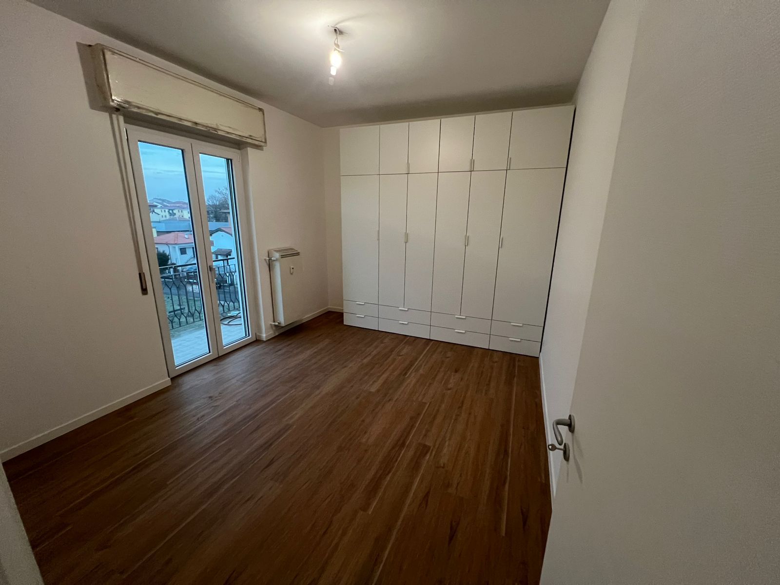 Camera da letto vuota con pavimento in legno, ante dell'armadio bianche e porta scorrevole in vetro che dà su un balcone.