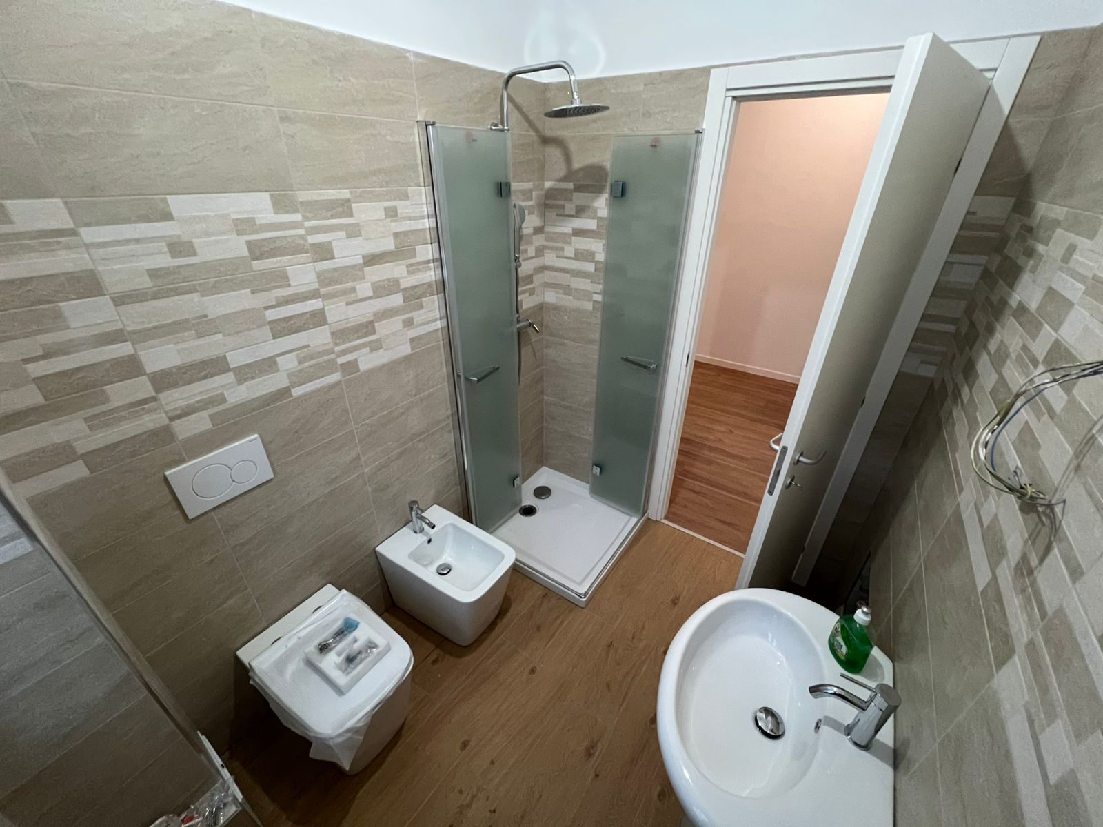 Piccolo bagno piastrellato con WC, bidet, box doccia, lavandino e porta aperta su una stanza con pavimento in legno.