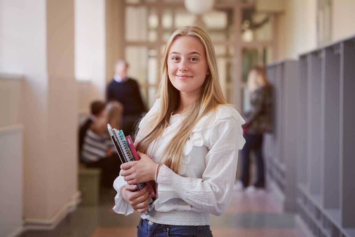 En ung kvinna står i en korridor med en bunt böcker i handen.
