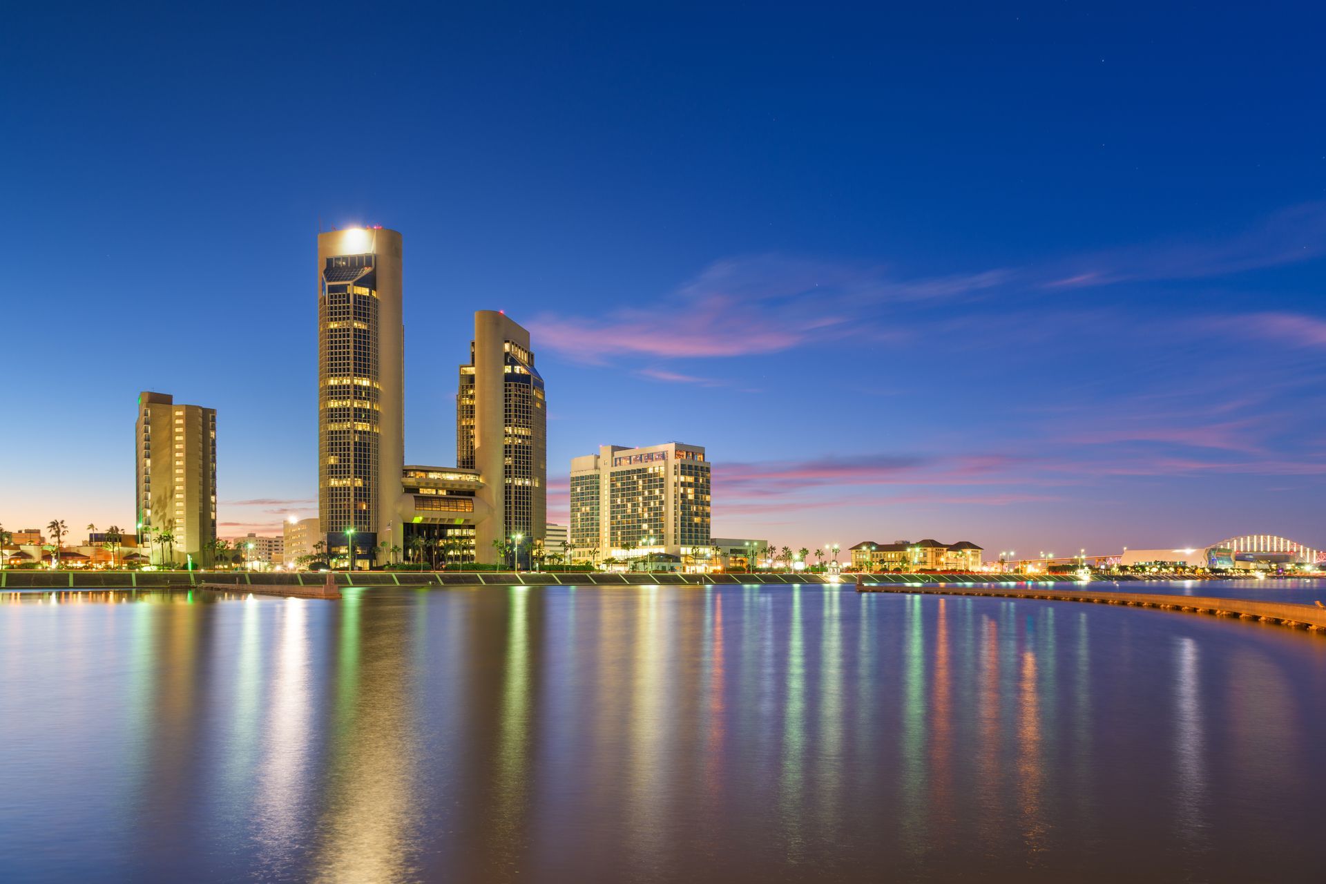 City skyline at dusk reflecting in calm water, with a blue sky and illuminated buildings.