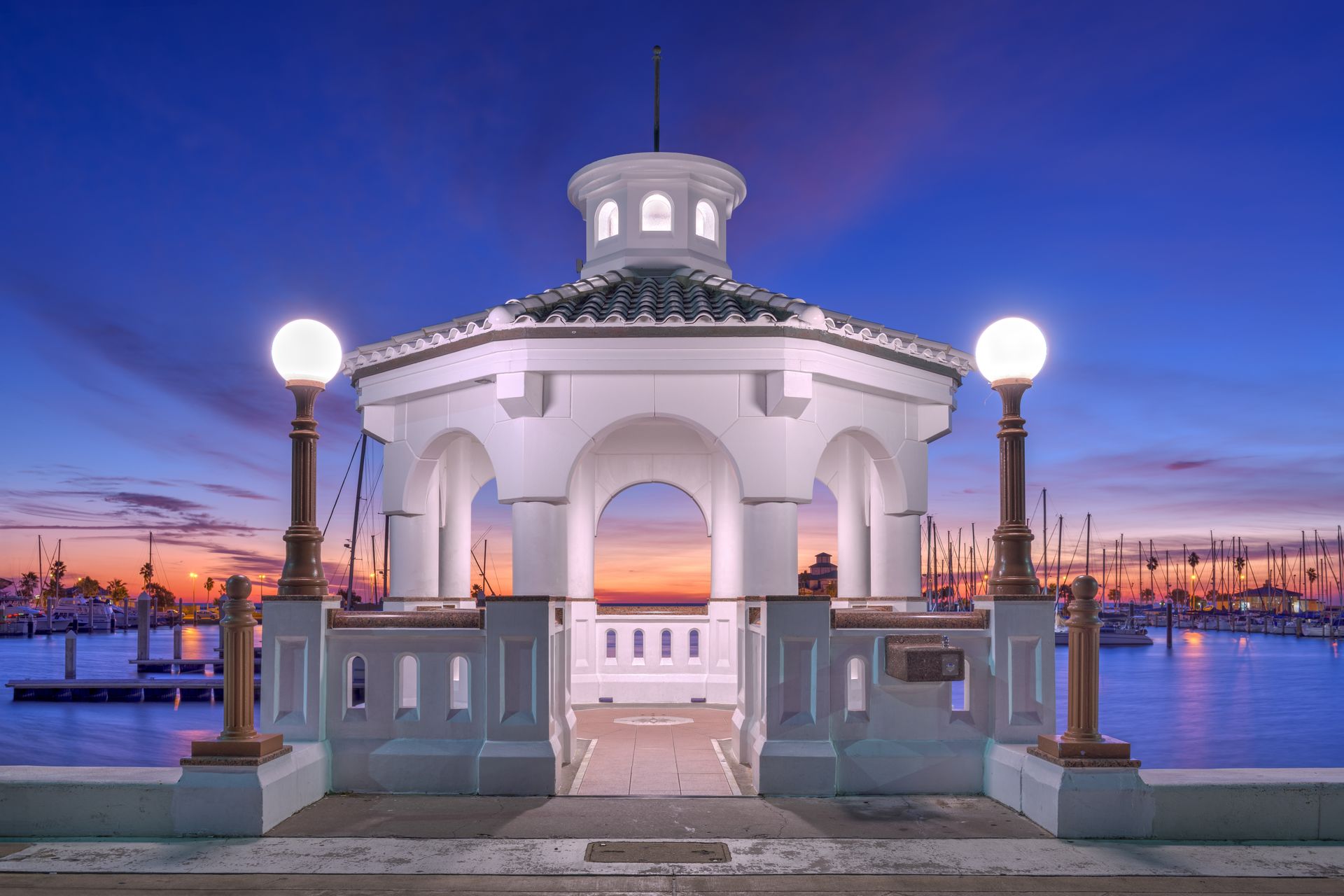 White gazebo with arched openings on a waterfront at dusk. Marina and sunset in the background.