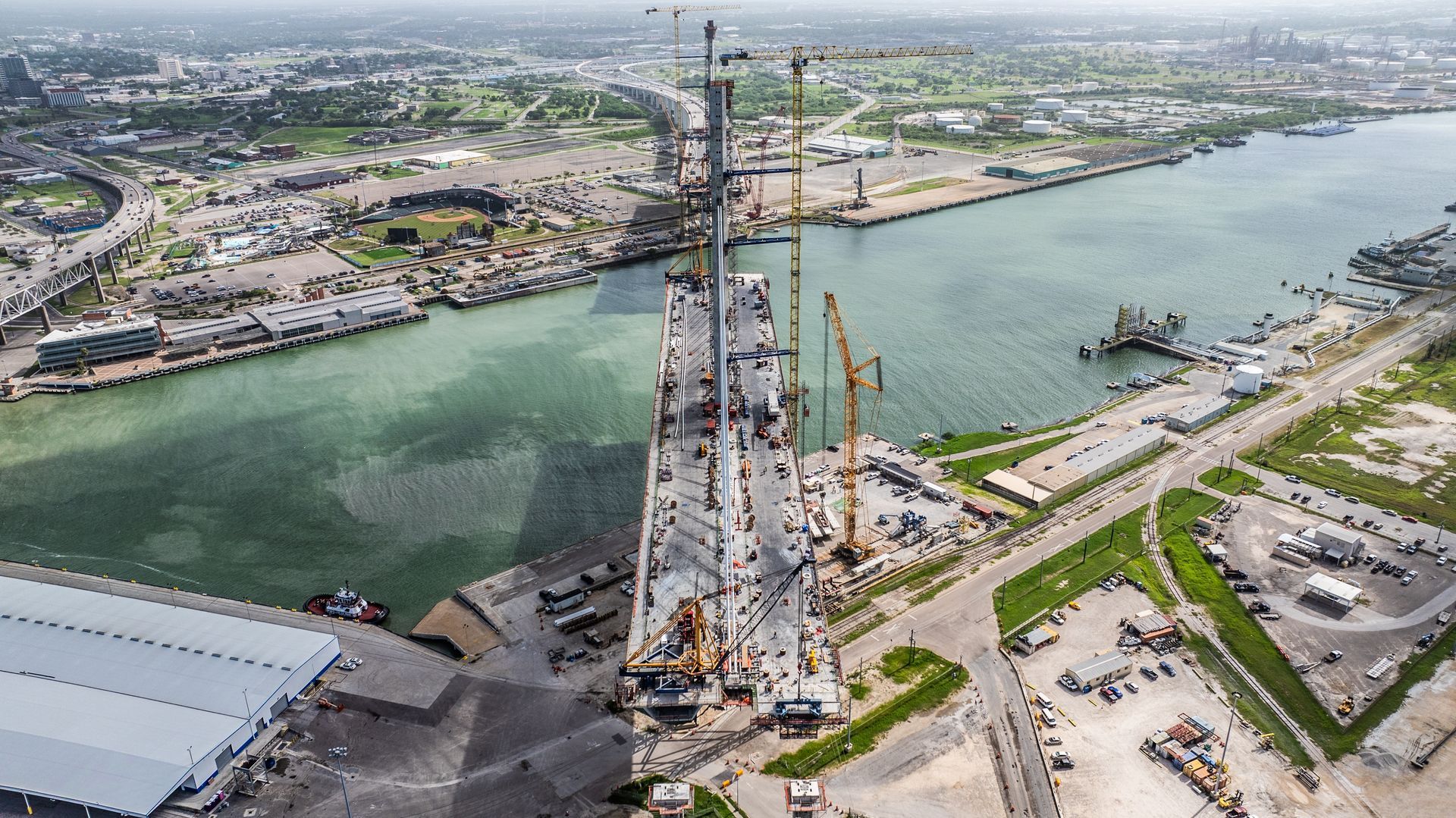 Aerial view of a bridge under construction, with cranes, a waterway, and surrounding buildings.