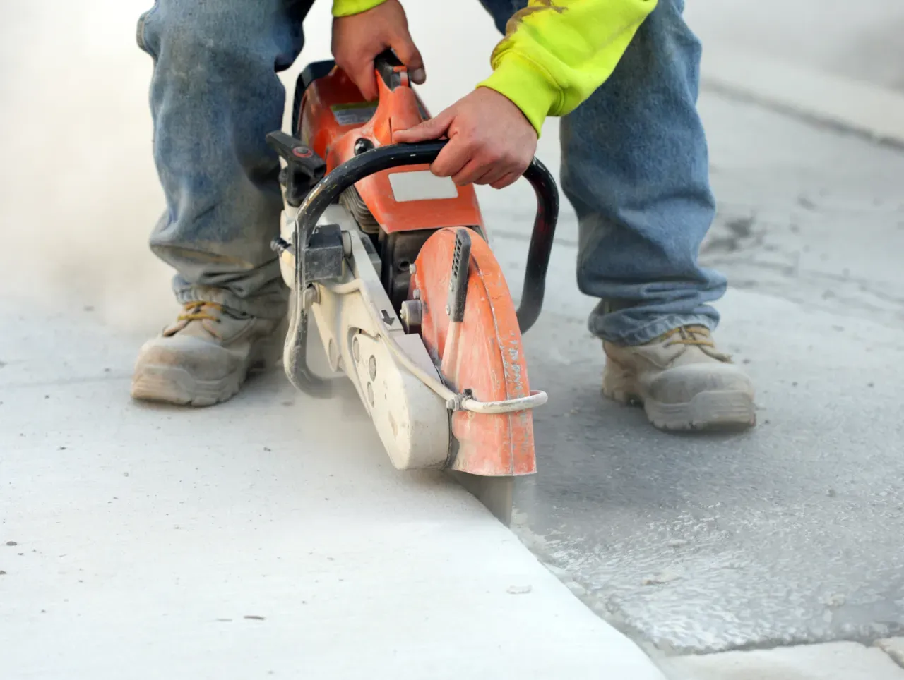 Construction worker sawing concrete pavement with an orange saw; dust flying.