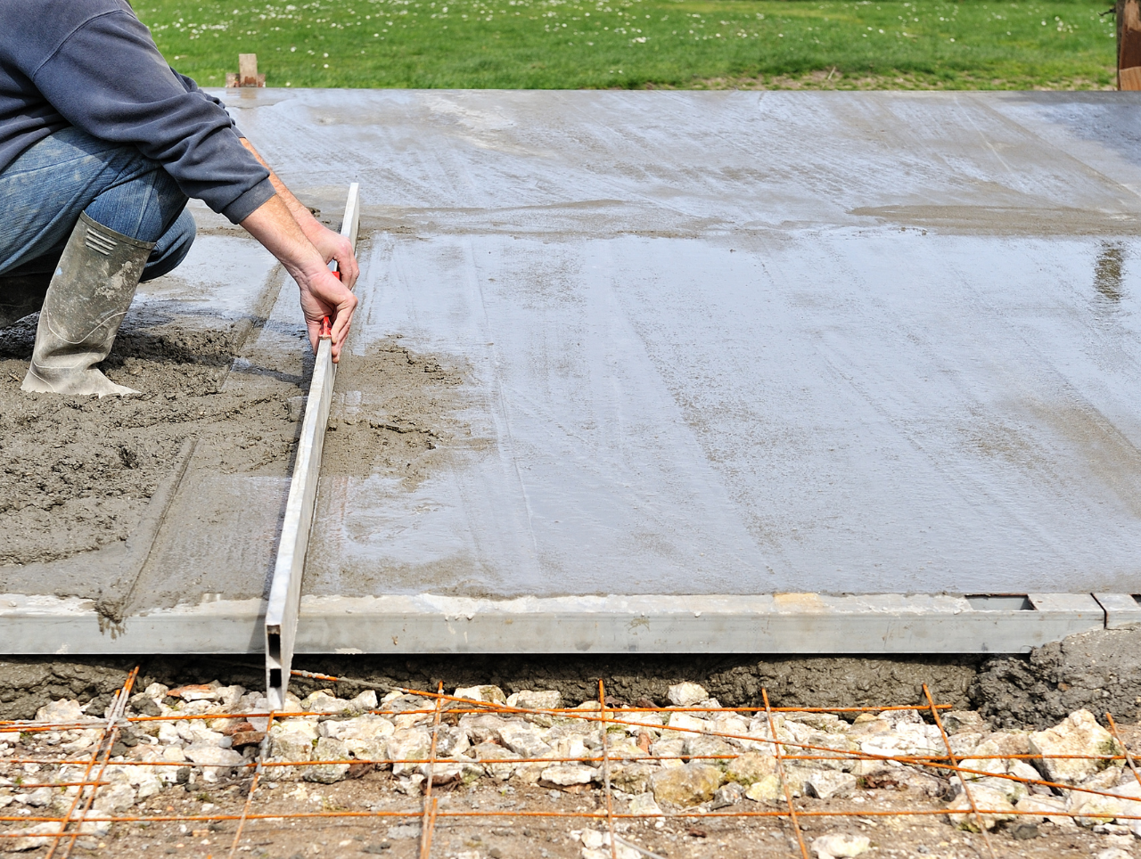 Person leveling wet concrete with a long tool, outdoor setting.