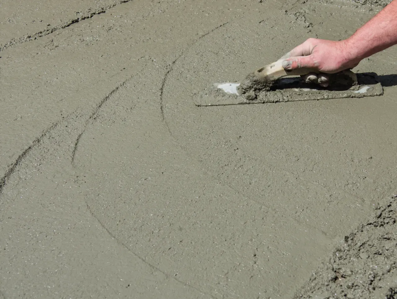 A person smoothing wet concrete with a trowel. Gray concrete, beige trowel, outdoors.
