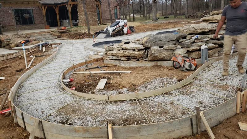 Construction of a curved concrete walkway with wooden formwork; a worker stands nearby.