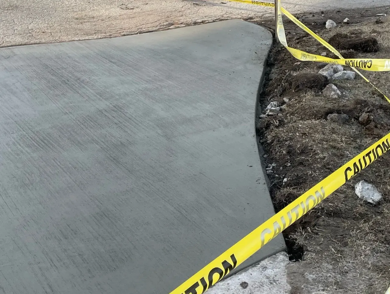 Newly poured concrete sidewalk with caution tape alongside dirt and stones.