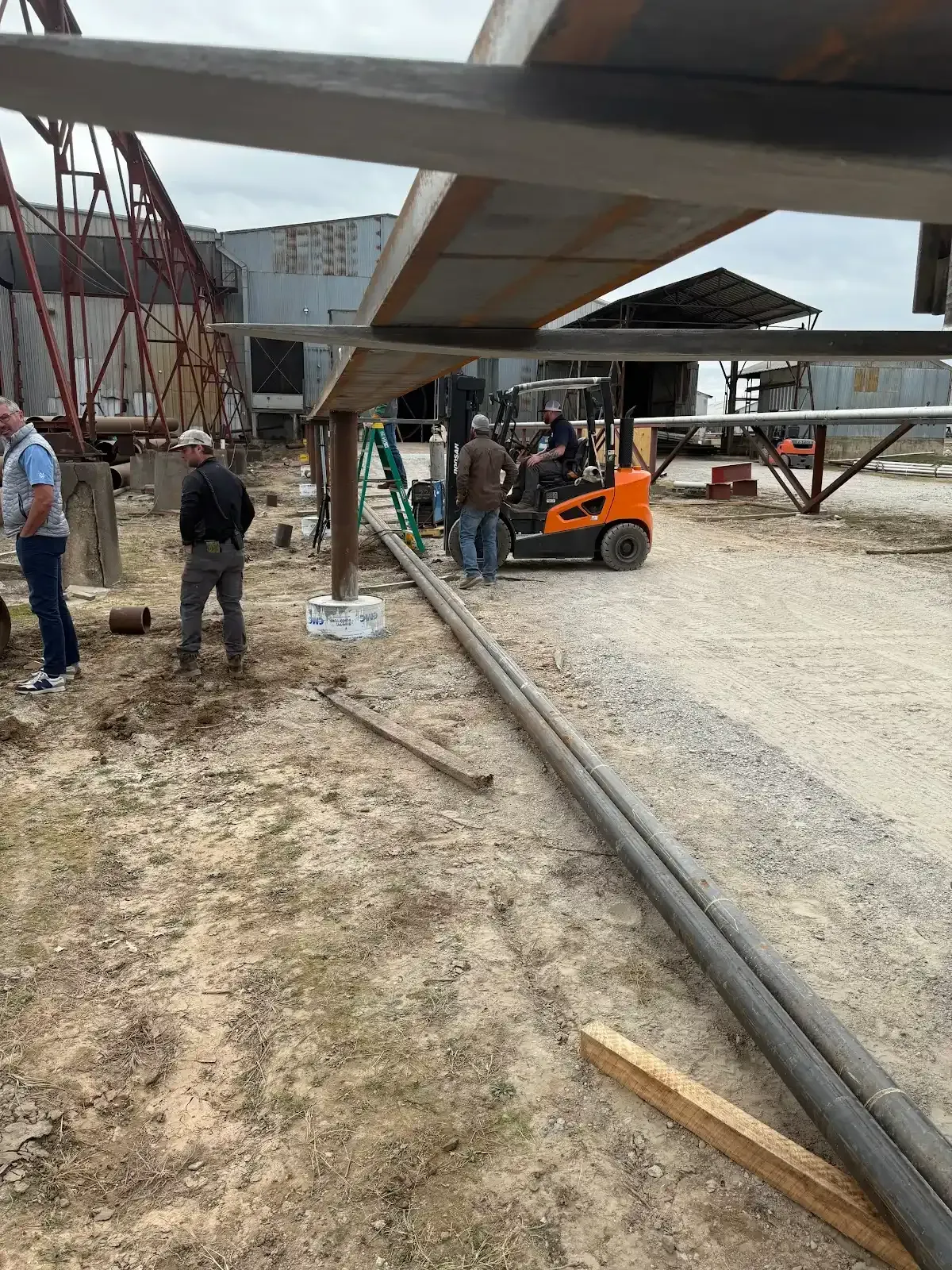 Workers constructing metal structure outdoors, using a forklift. Gray gravel ground and overcast sky.