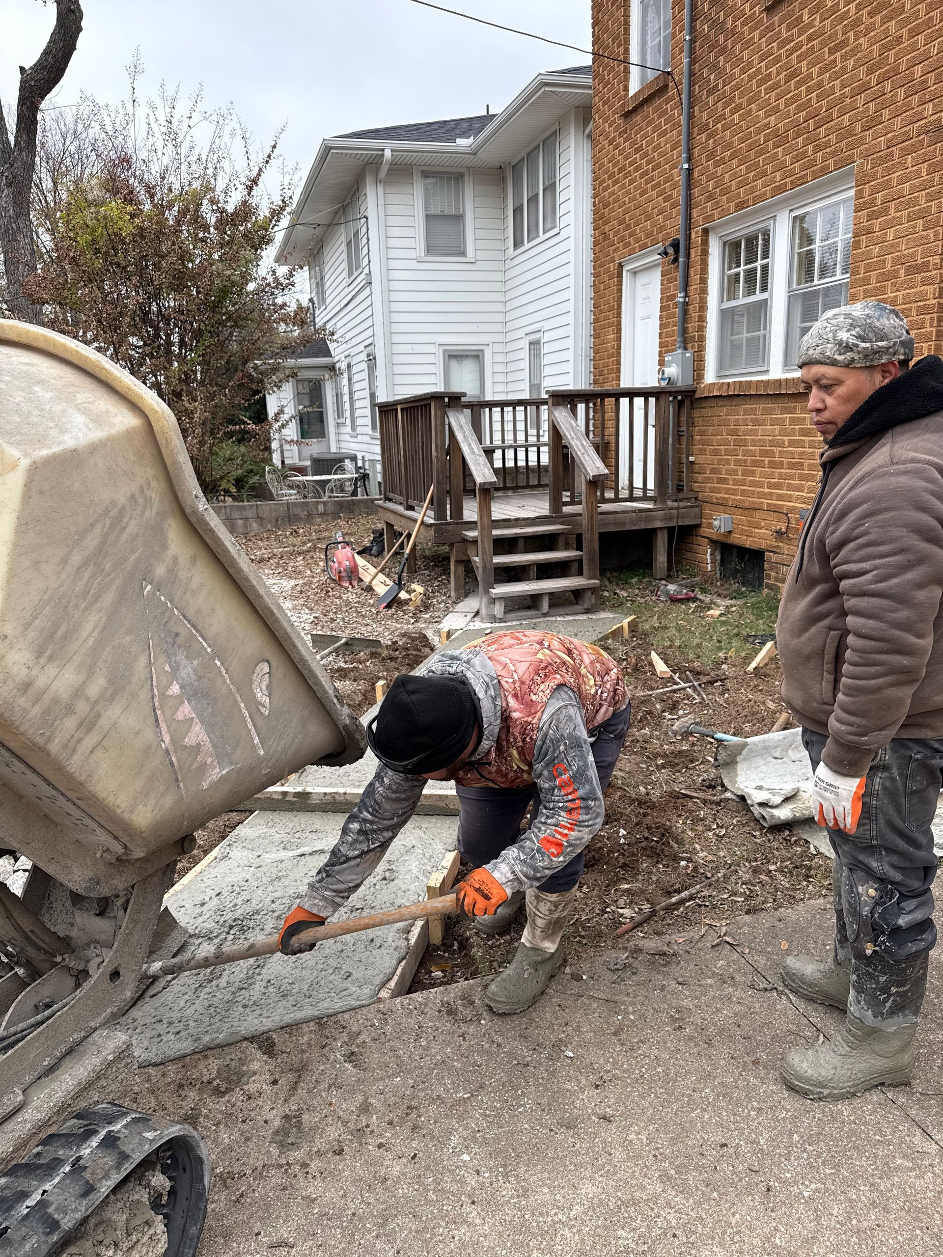 Two workers pouring concrete from a cart onto a sidewalk, near two-story buildings.