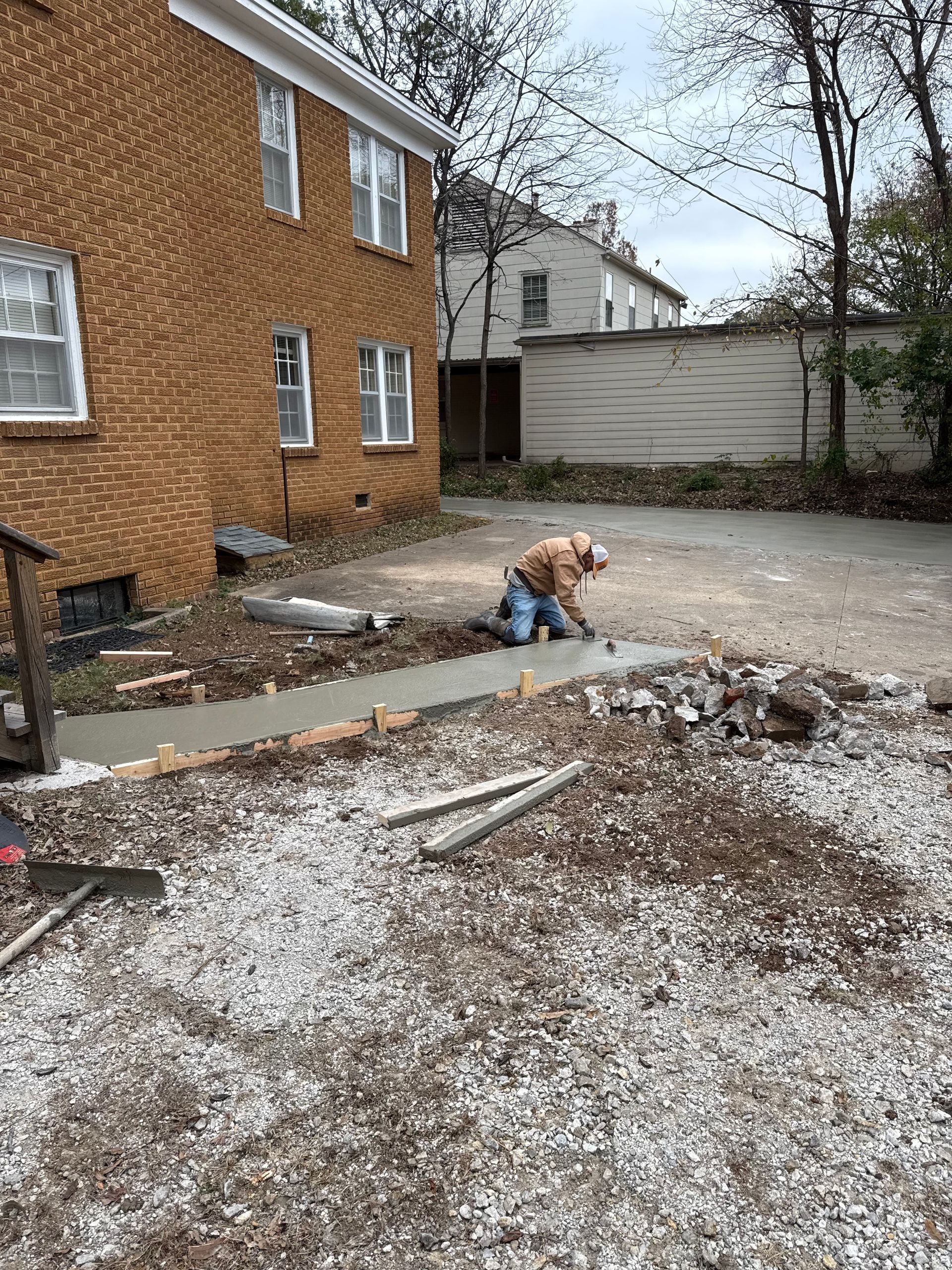 Man kneeling, smoothing wet concrete sidewalk alongside a brick building in an outdoor setting.