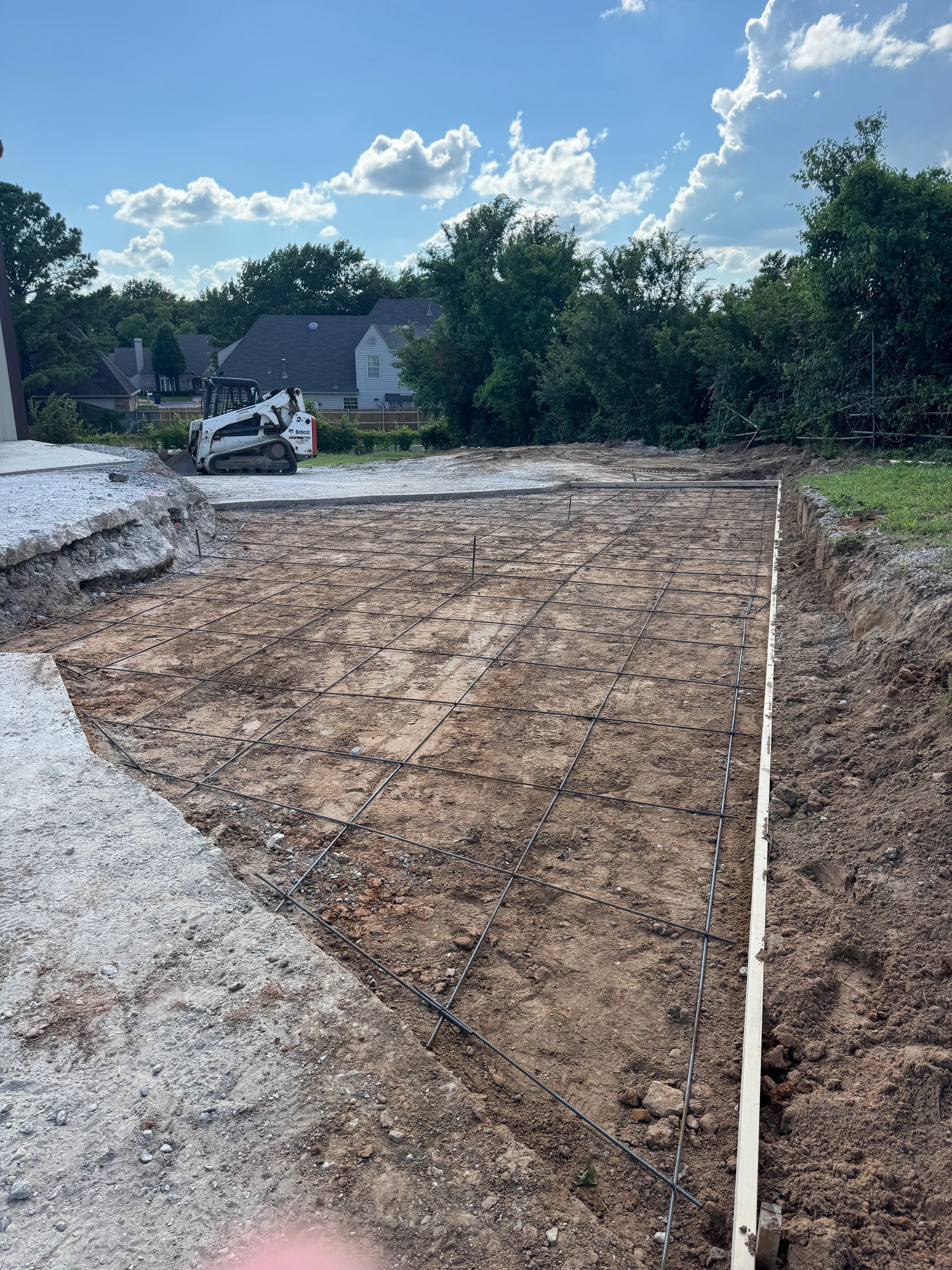 Construction site: earthworks, a Bobcat, and a gravel foundation. Green grass, trees, and blue sky in the background.