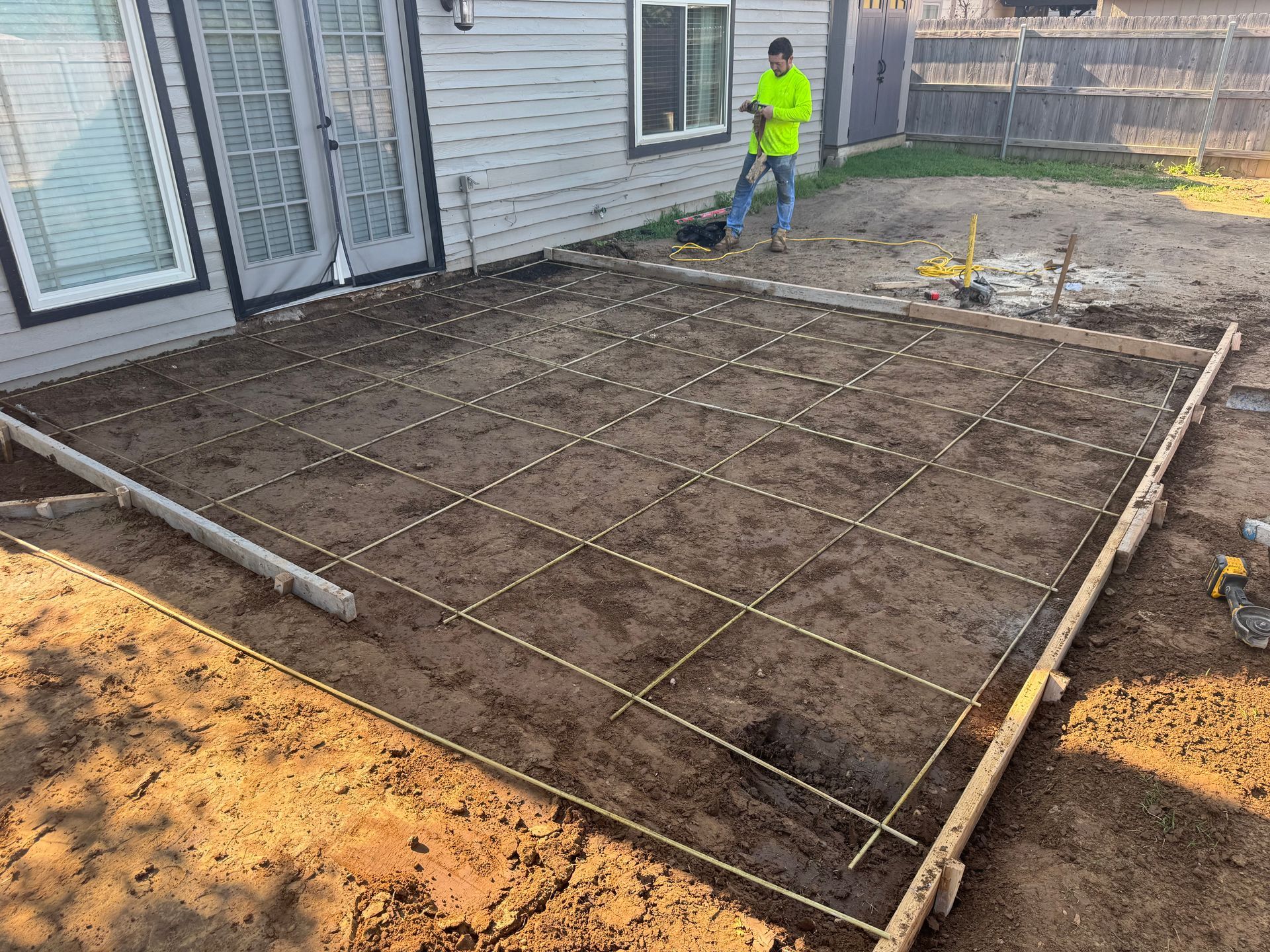 Construction of a concrete patio: Man working, rebar grid laid out in prepared ground. Gray house in background.