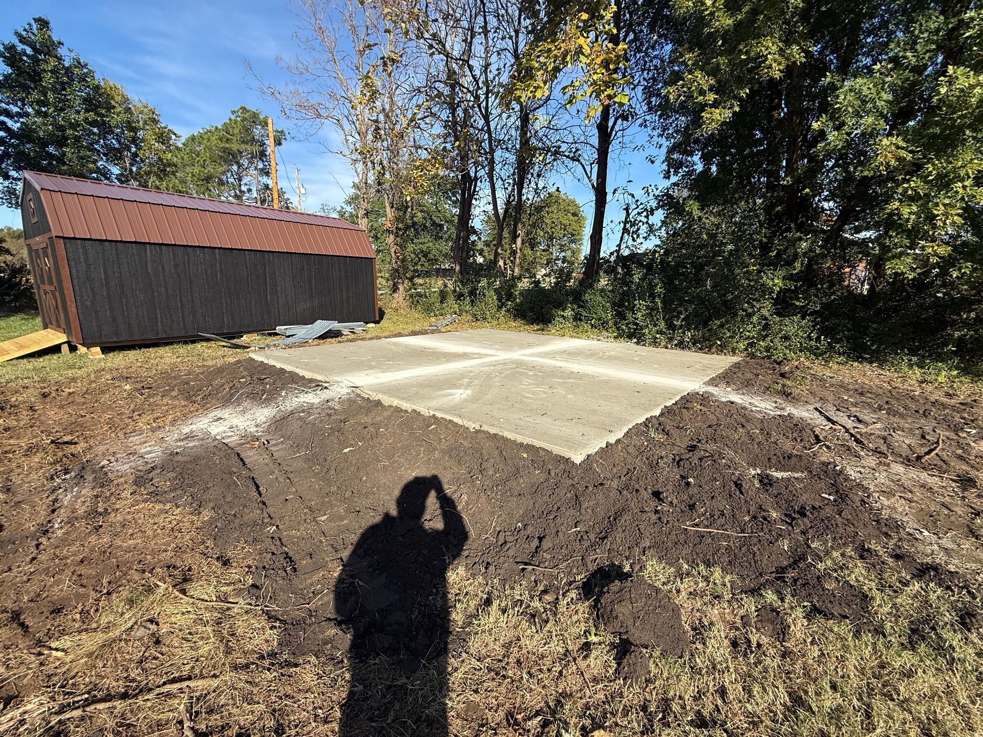 A person's shadow on dirt in front of a concrete foundation with a shed in the background.