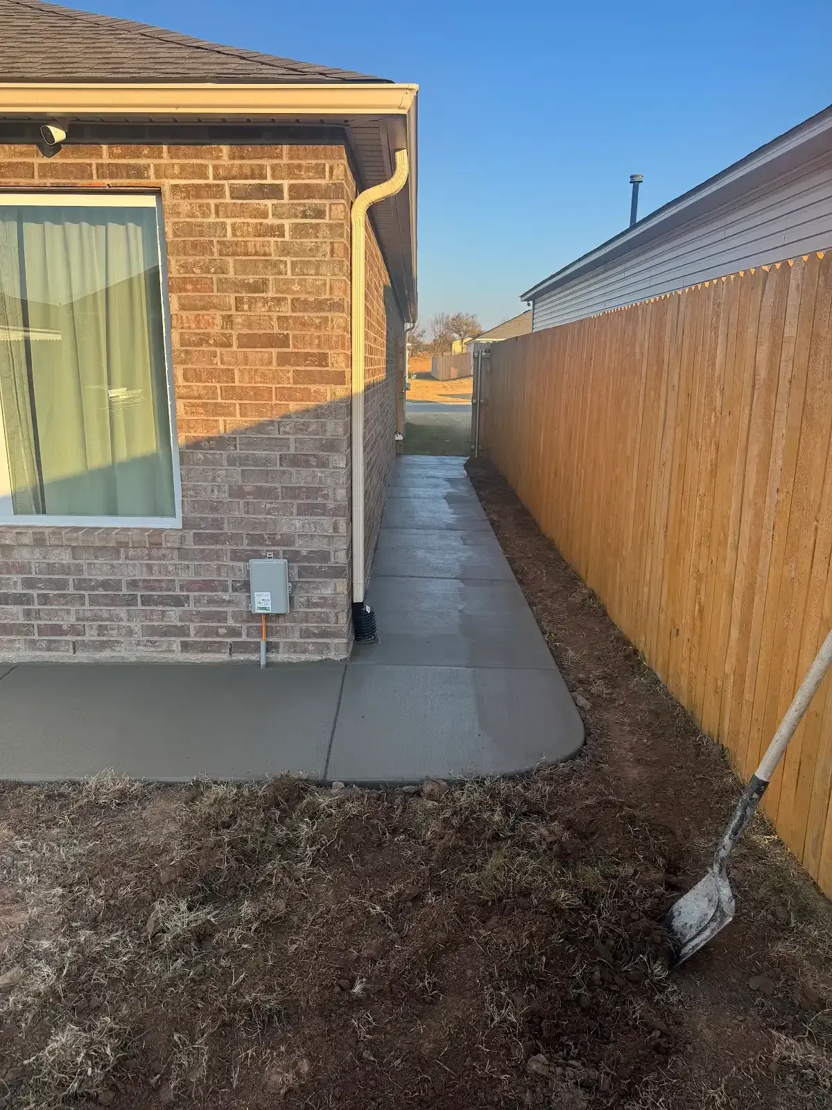 Concrete pathway alongside a brick house and wooden fence. A shovel rests against the fence on the right.
