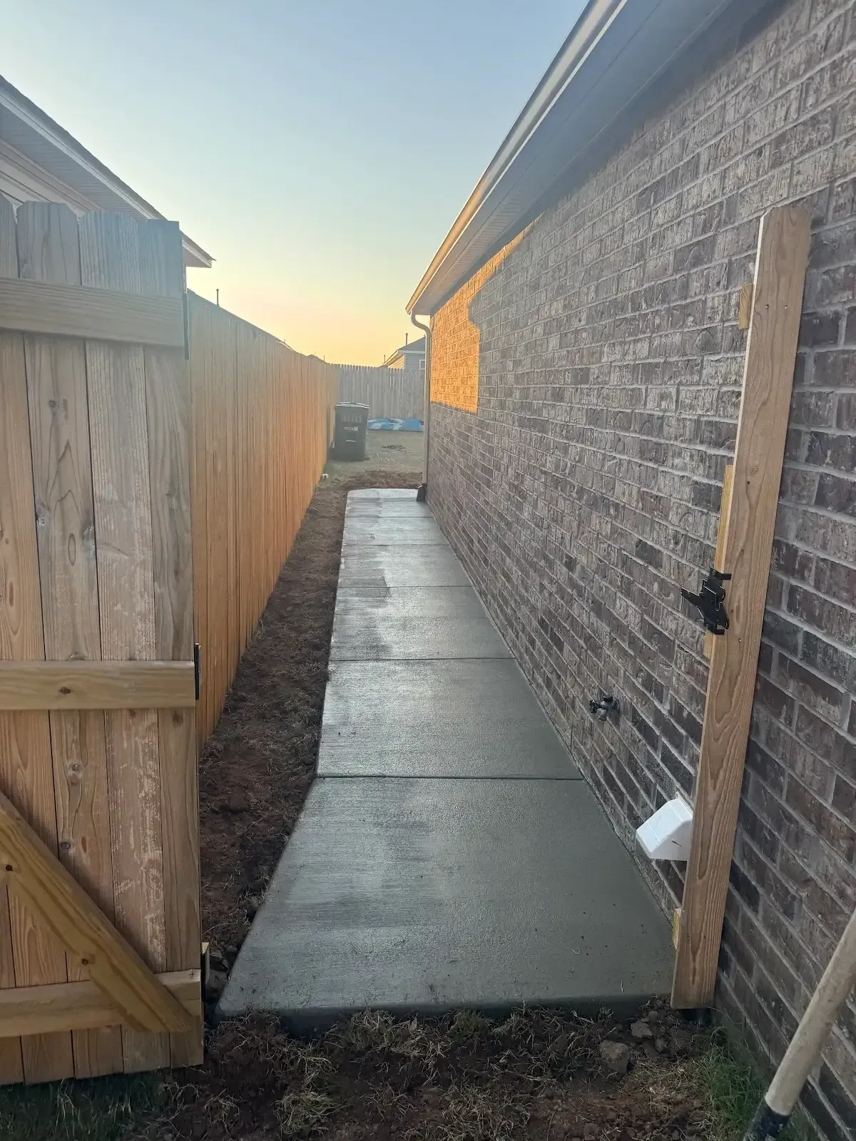 Concrete walkway between brick wall and wooden fence, leading to a gate.