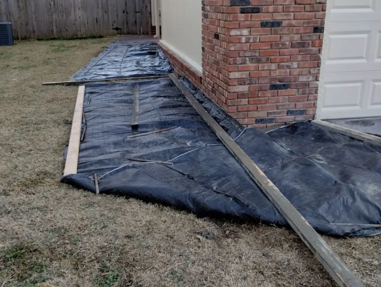 Black tarp laid on grass near a red brick building and garage, with wooden planks.