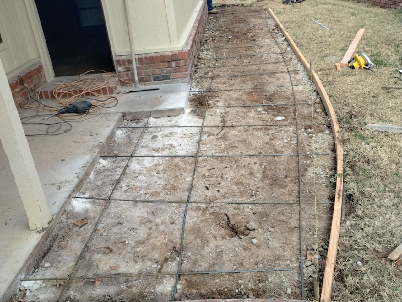 Concrete pathway under construction with rebar grid. Wooden form curves along the edge, with tools nearby on grass.