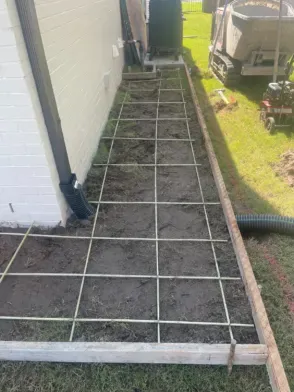 A concrete pathway under construction, with rebar grid and wooden frame, beside a house and lawn.