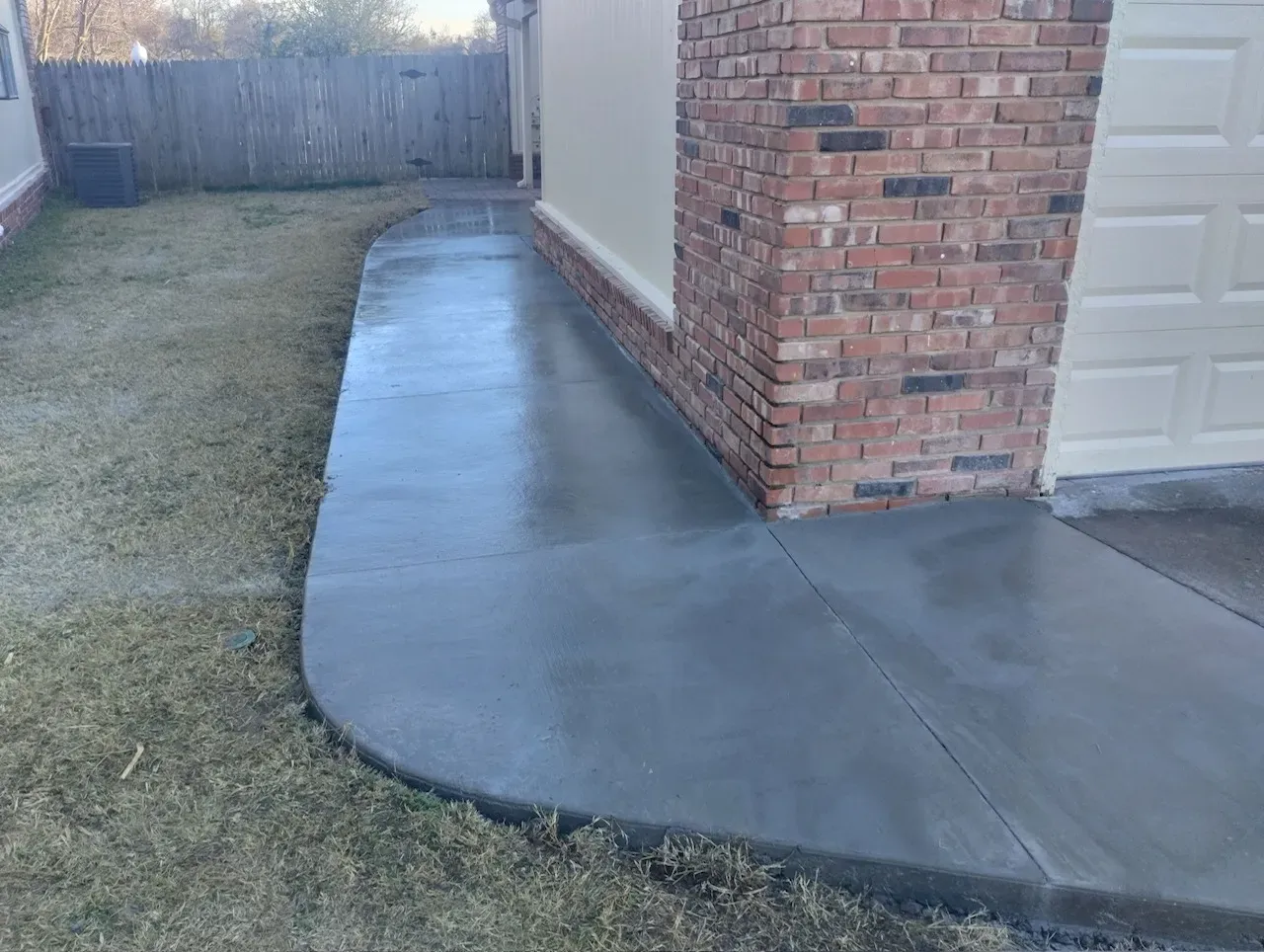 Freshly poured concrete walkway next to a brick wall and garage door; curving path through yard.
