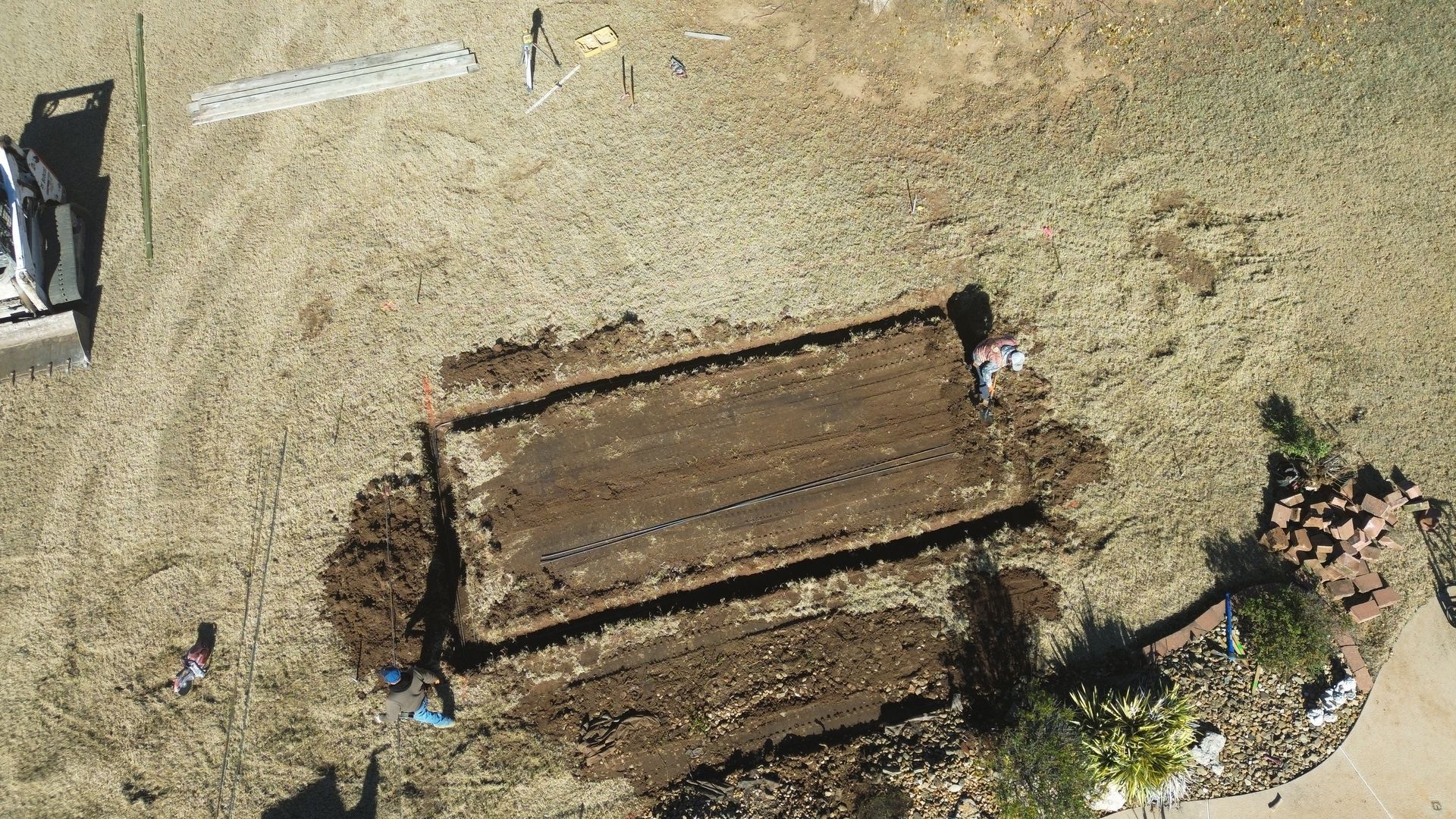 Overhead view of a rectangular garden bed dug in dry grass. Soil is tilled, with two people working nearby.