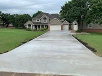 A large, newly poured concrete driveway leads to a two-story house with a green lawn and overcast sky.
