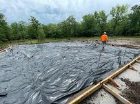 Person in orange shirt walks on a large tarp-covered foundation in a wooded area.