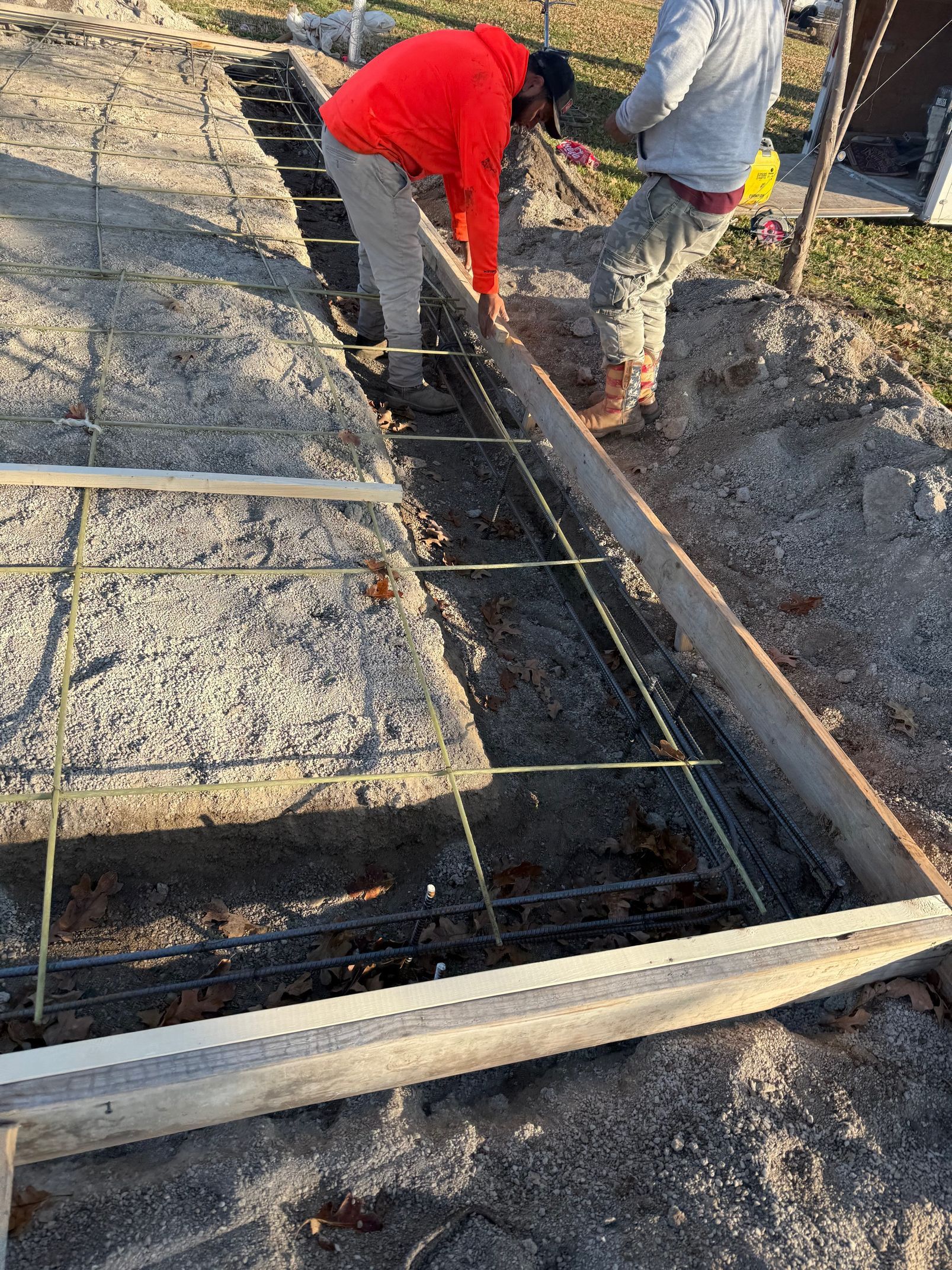 Two construction workers setting rebar in a concrete form. One wears a red shirt.