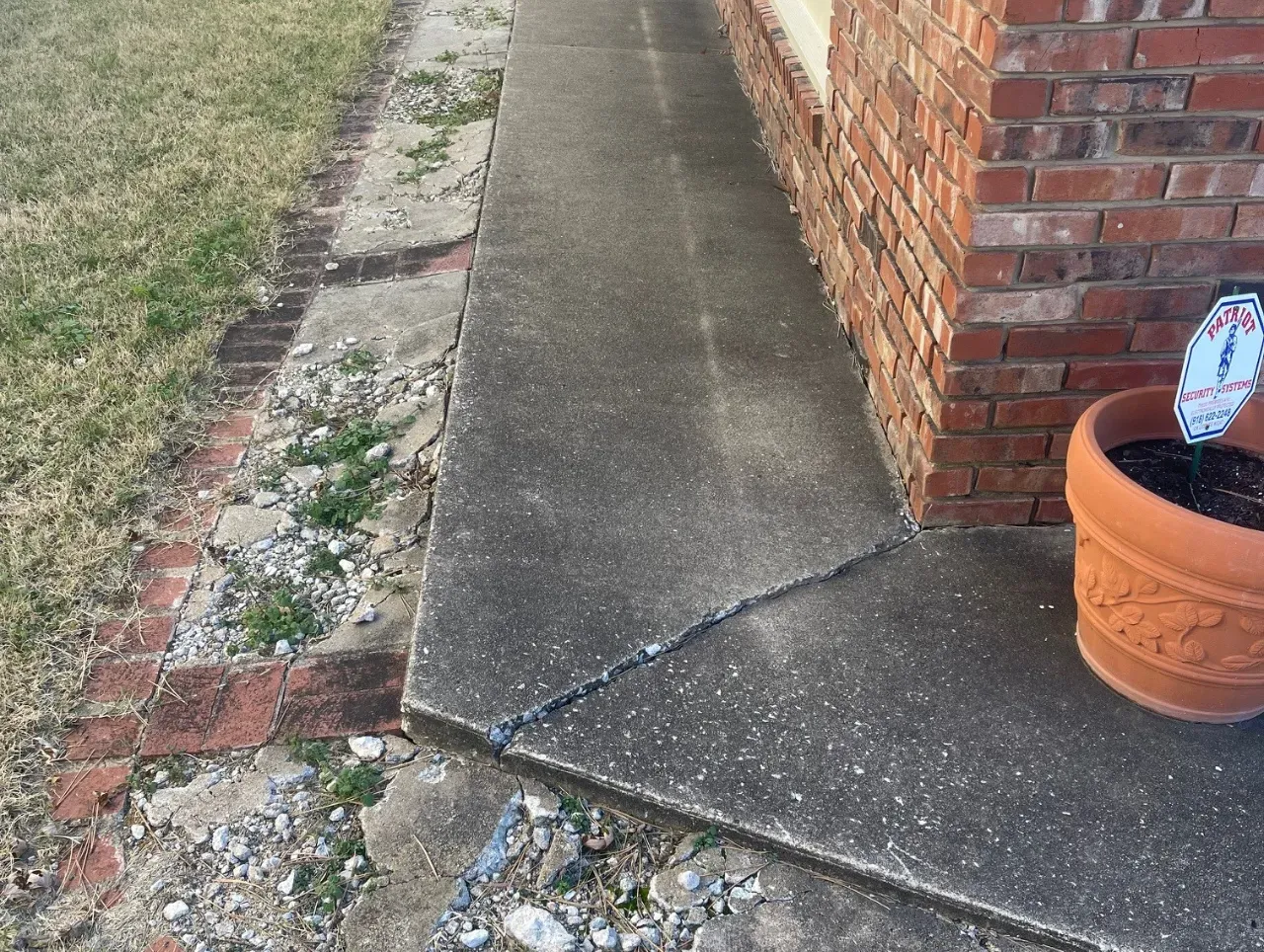 Cracked concrete sidewalk next to a brick wall and a flower pot.