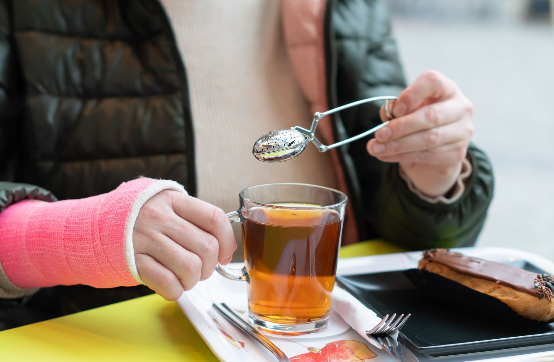 Eine Frau mit einem Gipsarm sitzt mit einer Tasse Tee und einem Löffel an einem Tisch. Nach einer Fraktur Ergotherapie