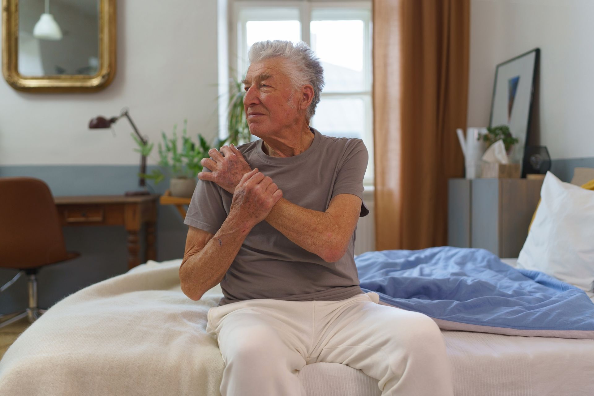 Ein älterer Mann sitzt mit den Händen auf den Armen auf einem Bett. Multriple Sklerose Ergotherapie