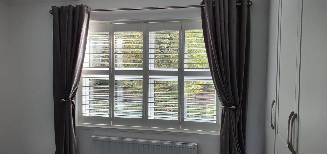 A bedroom with a window with shutters and curtains.