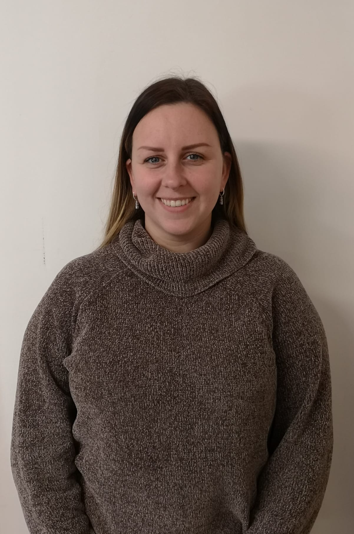 A woman in a brown sweater is smiling and standing in front of a white wall.