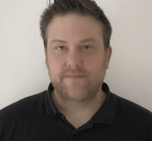 A man with a beard is wearing a black shirt and looking at the camera.