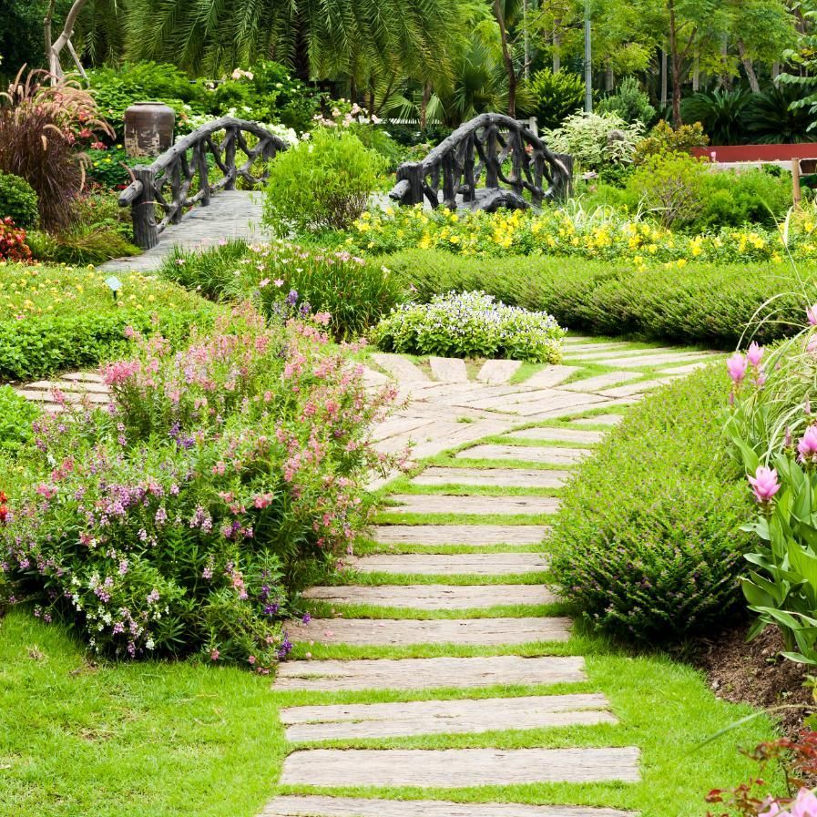 Stone pathway through a lush garden, leading to a bridge with green foliage and colorful flowers.