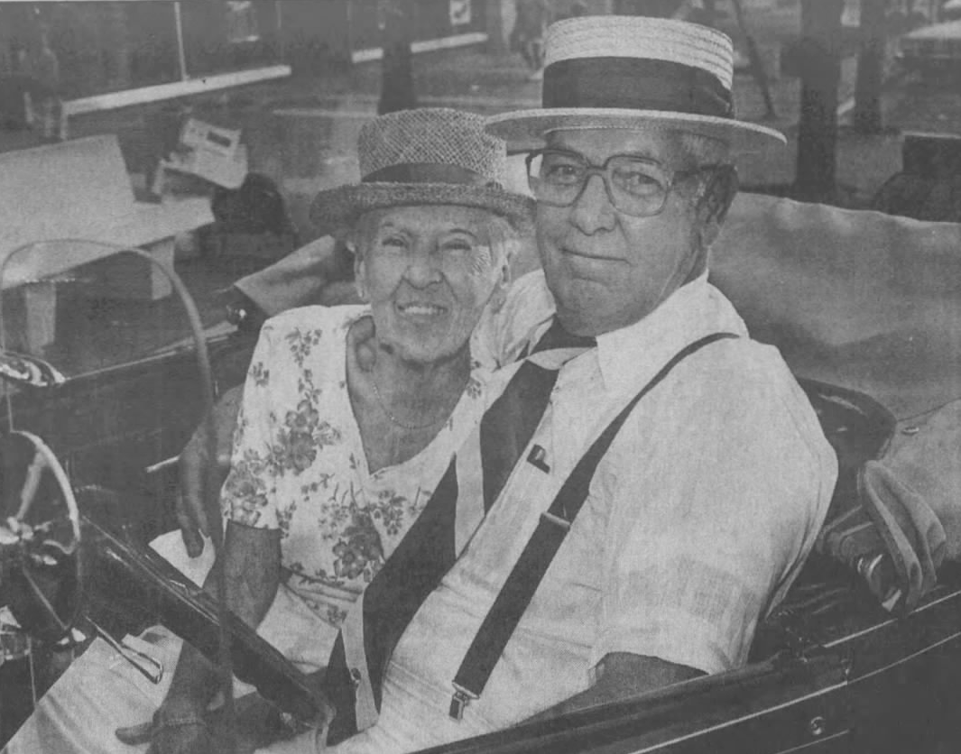 Elderly couple in a classic car, both wearing hats and smiling. Man driving, woman seated beside him.