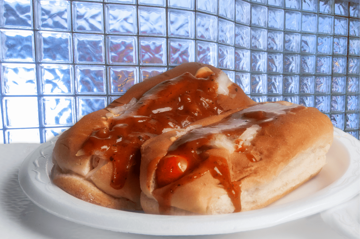 Two Chris' Famous chili dogs on a white plate; glossy sauce drips down the buns, against a textured blue backdrop.