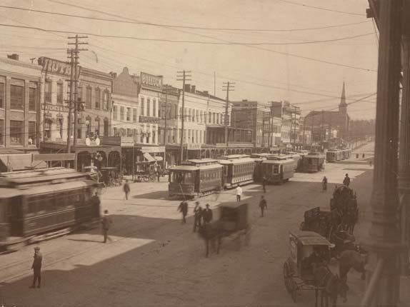 A black and white photo of a city street with horse drawn carriages.