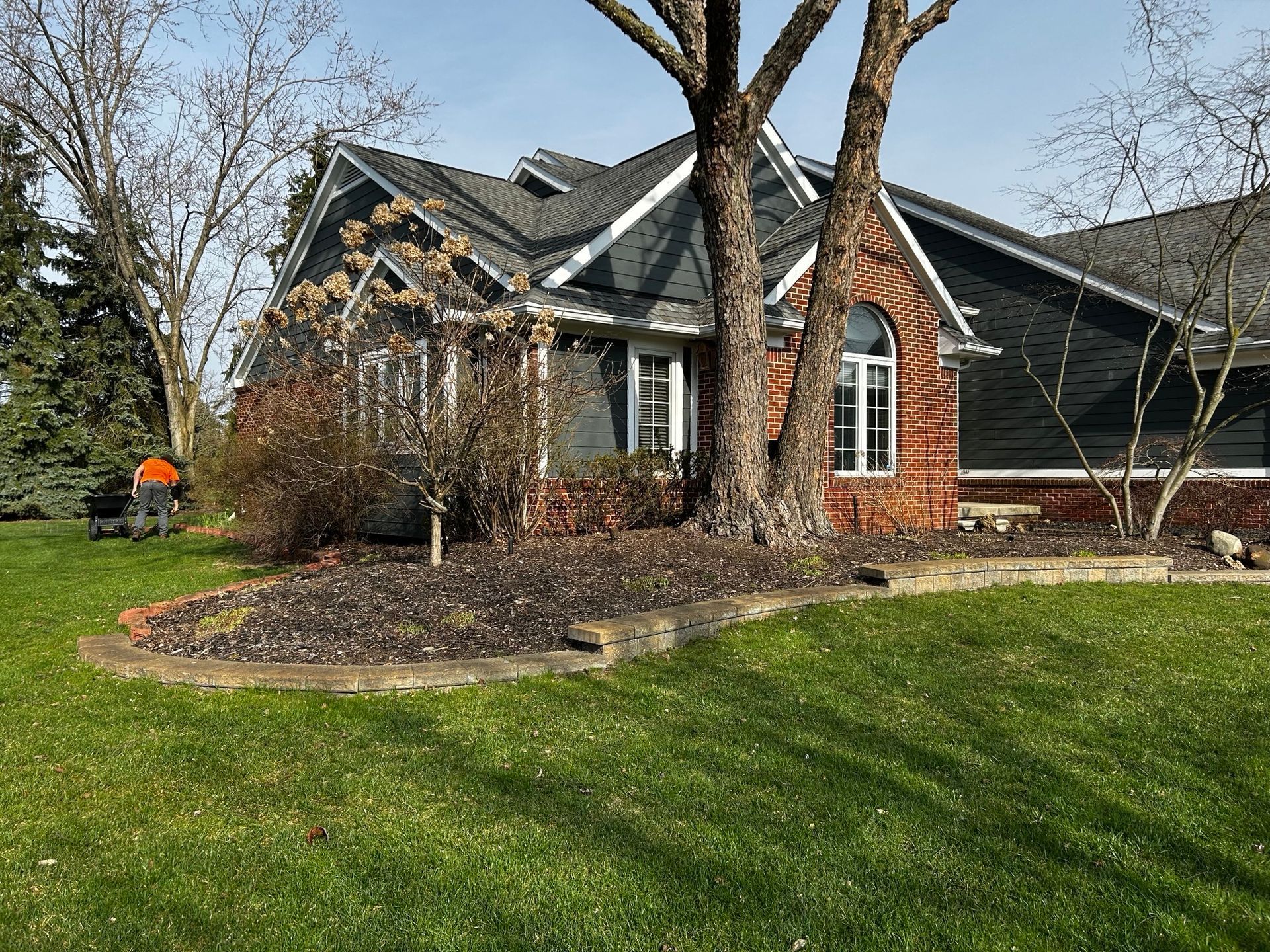 House with blue siding, red brick, and a tree in front. A lawn mower is visible on the left.