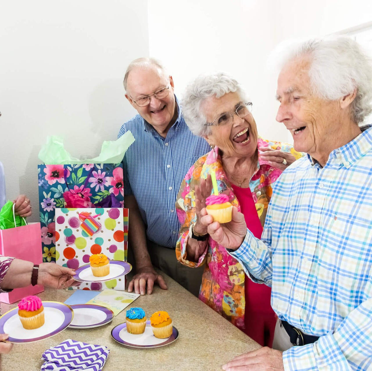 Group of vibrant independent living senior residents celebrating a friends birthday with cupcakes and gifts