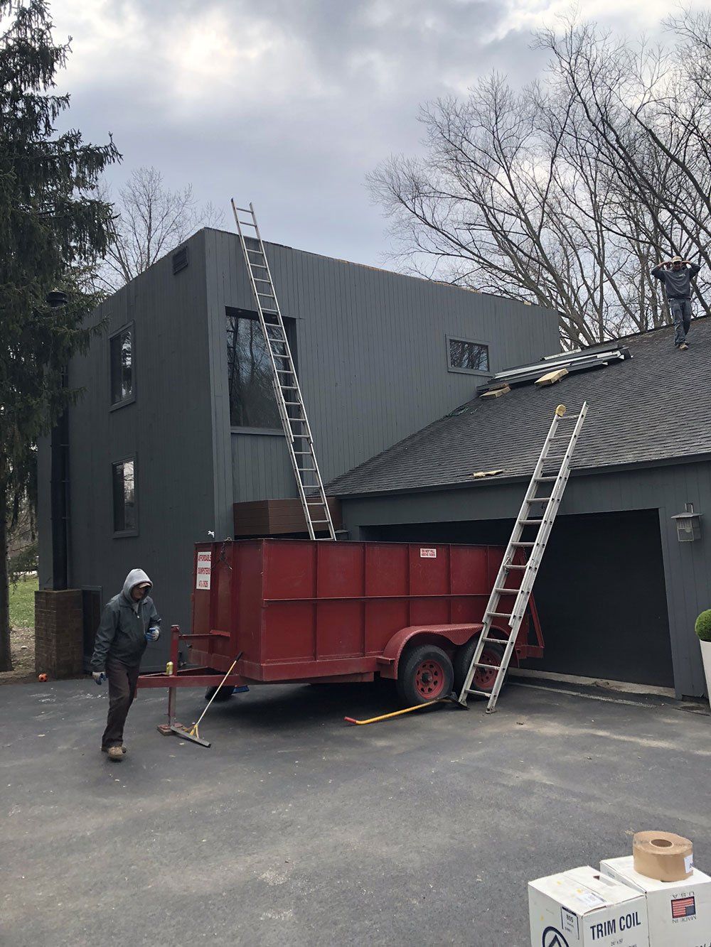 A man is standing in front of a house with a red trailer and ladder.