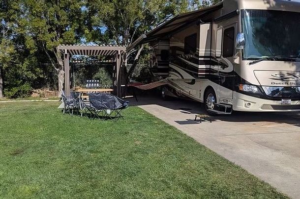 A beige recreational vehicle with an extended awning parked next to a pergola on a grassy lot with outdoor chairs.