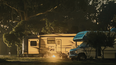 A camper trailer and a motorhome parked in a wooded area under the warm, golden light of sunset.
