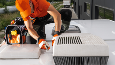 A technician in an orange uniform and work gloves works on an air conditioning unit on top of a white vehicle roof.