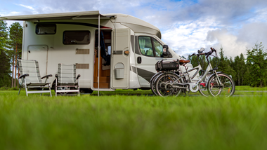 A white camper van with an extended awning parked in a grassy field next to two bicycles and two outdoor chairs.