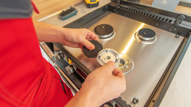 A person in a red uniform installs a gas burner cap onto a stainless steel stove top.