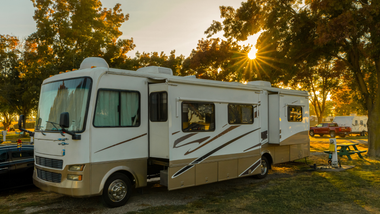 A white and beige Class A motorhome parked at a campsite during a sunny sunset among trees.
