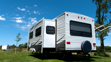 A white travel trailer with a slide-out section sits on a grassy field under a sunny blue sky.