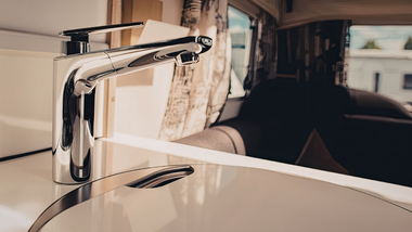 A sleek chrome kitchen faucet sits on a white countertop inside a modern camper or RV with soft-focus interior seating.