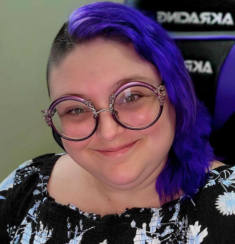 a white woman with purple hair smiles at the camera. She's wearing purple-framed glasses and a black shirt with flowers.